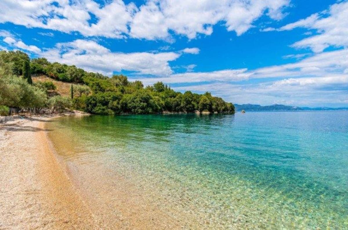 Ευρεία λψη μιας βοτσαλωτής παραλίας με καθαρά τιρκουάζ νερά που οδηγούν σε μια δασώδη ακτογραμμή κάτω από μπλε συννεφιασμένο ουρανό.