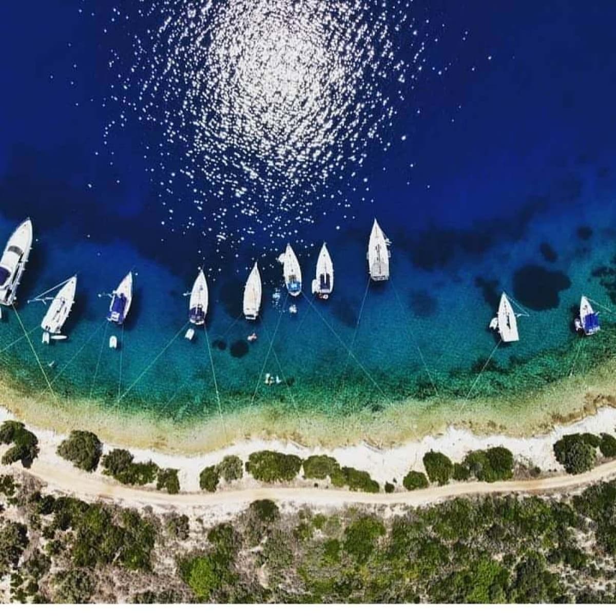 Αεροφωτογραφία δείχνει ιστιοφόρα αγκυροβολημένα κοντά σε βραχώδη ακτή με γαλαζοπράσινα νερά.
