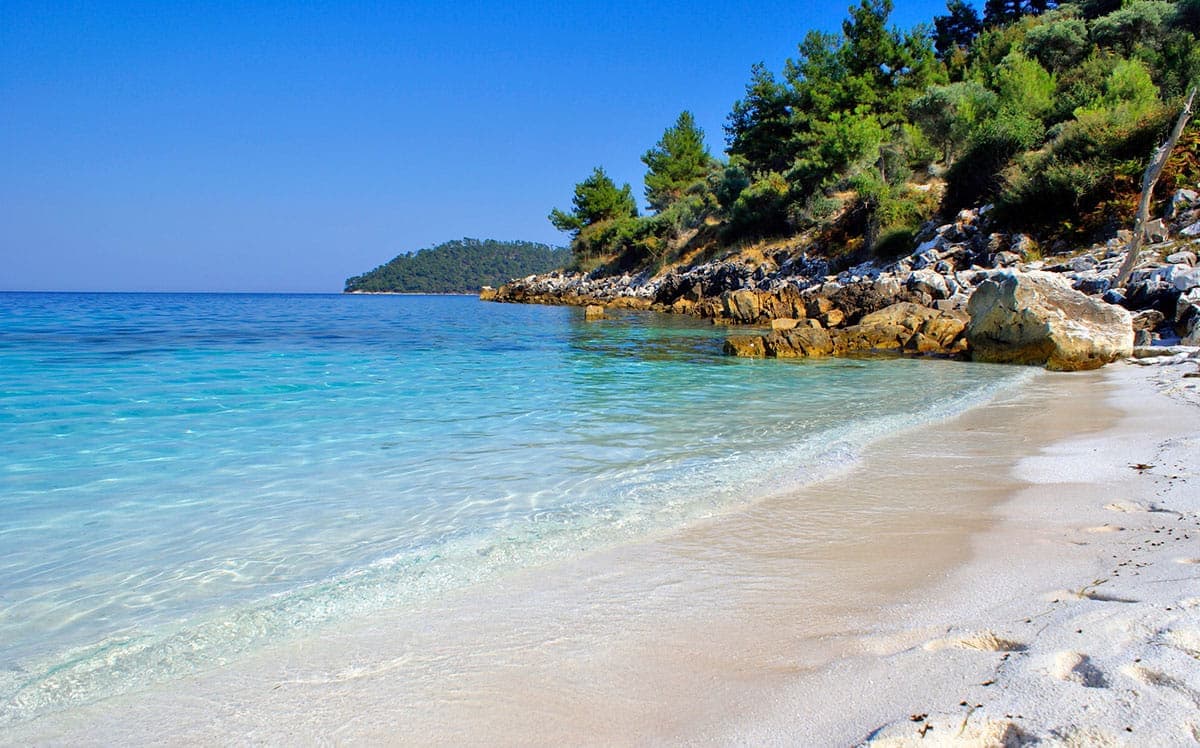 Παραλία με λευκή άμμο, γαλαζοπράσινα νερά και δέντρα.