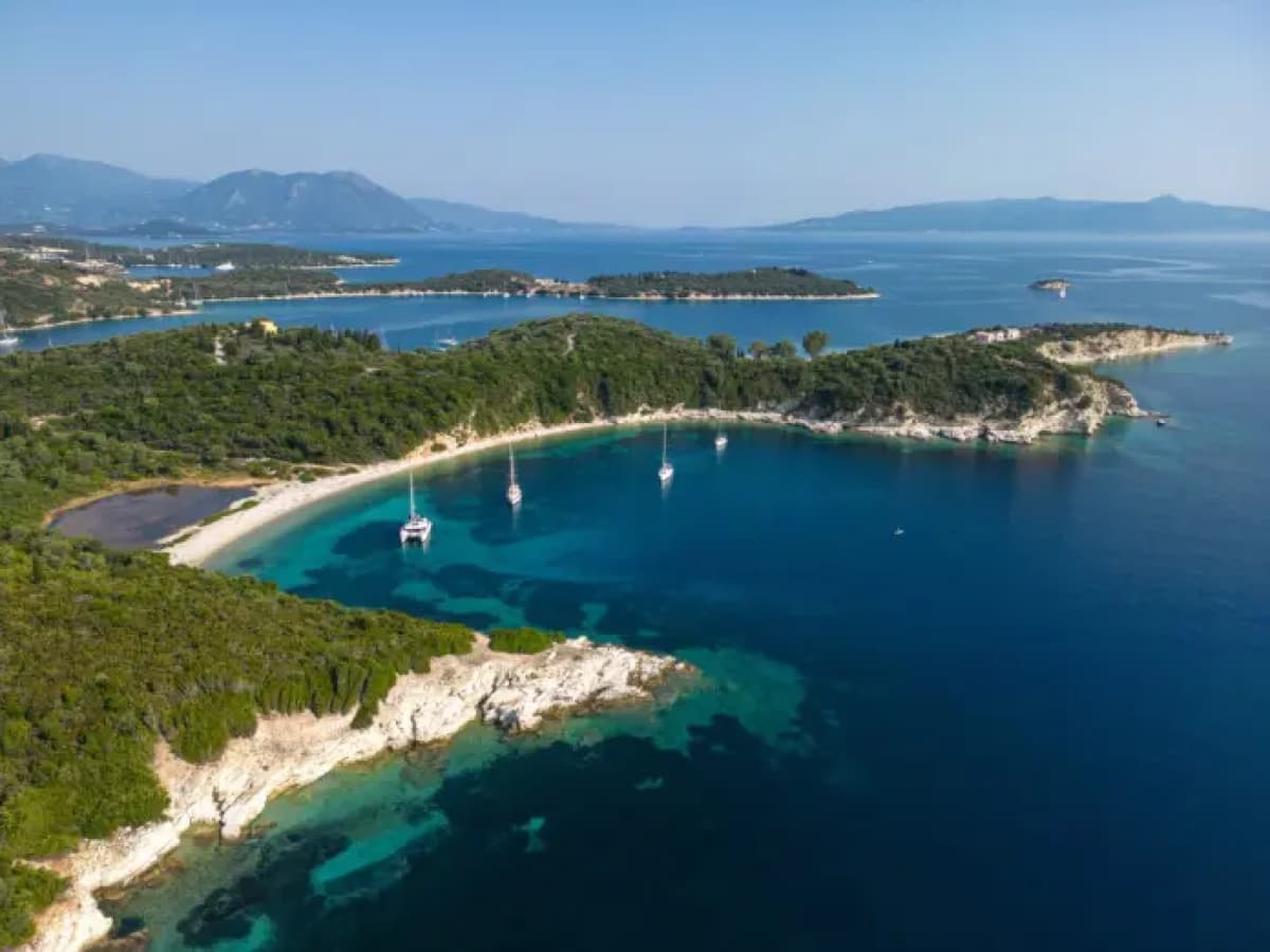 Εναέρια άποψη παράκτιας περιοχής με δάσος, παραλία, βράχους και ιστιοφόρα στα γαλαζοπράσινα νερά.