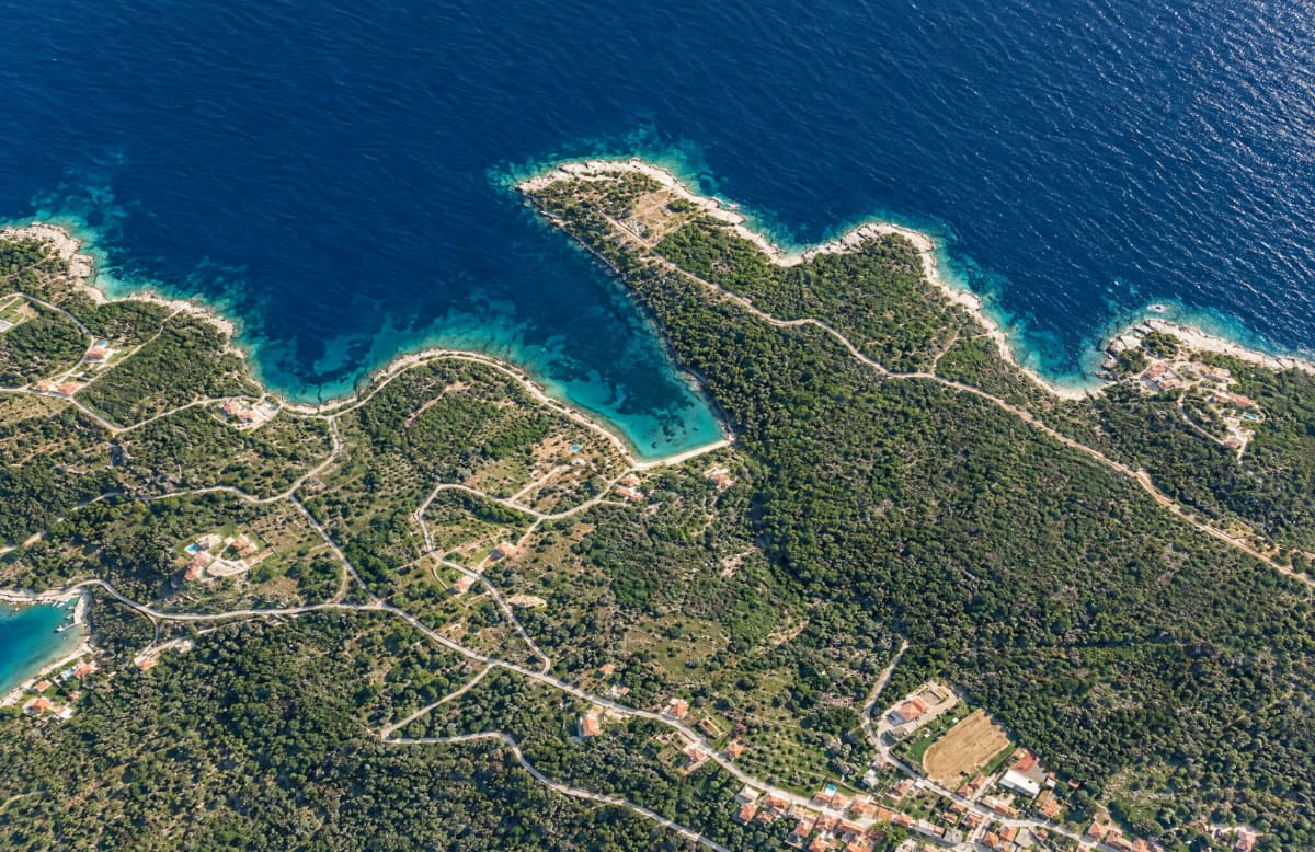 Εναέρια άποψη μιας δασώδους ακτογραμμής με ελικοειδείς δρόμους, κτίρια και γαλάζια θάλασσα.