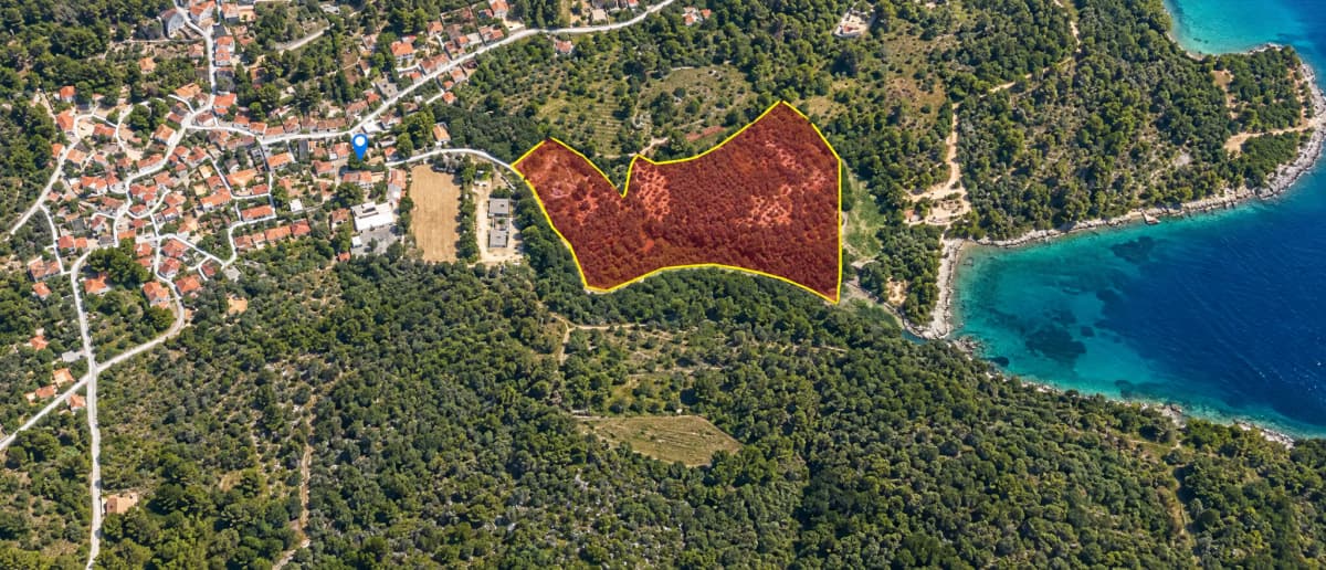 Εναέρια άποψη δασώδους παράκτιου τοπίου με επισημασμένο οικόπεδο κοντά σε χωριό και θάλασσα.