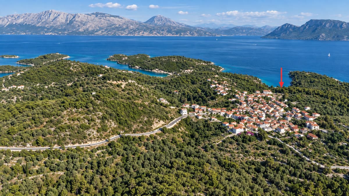 Εναέρια άποψη ενός πυκνού πράσινου δάσους με ένα χωριό με λευκά σπίτια και πορτοκαλί στέγες, με θέα στη θάλασσα και τα βουνά στο βάθος.