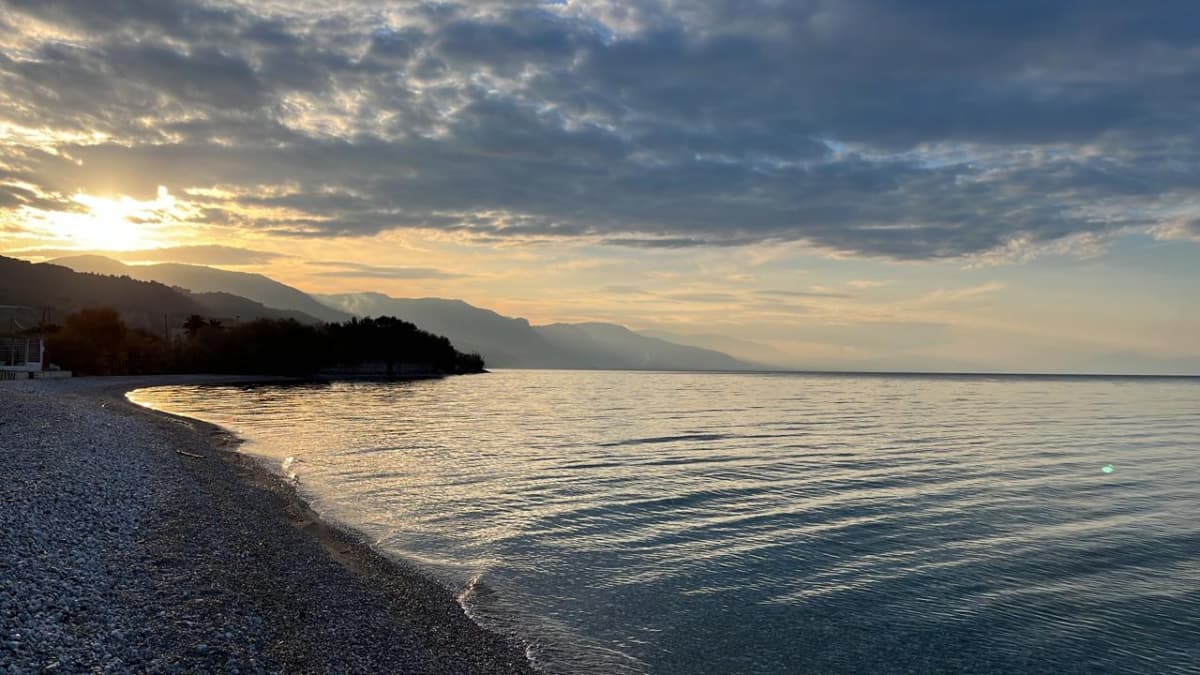 Ευρεία θέα μιας πετρώδους παραλίας και ήρεμης θάλασσας κάτω από συννεφιασμένο ουρανό κατά το ηλιοβασίλεμα, με βουνά στο βάθος.