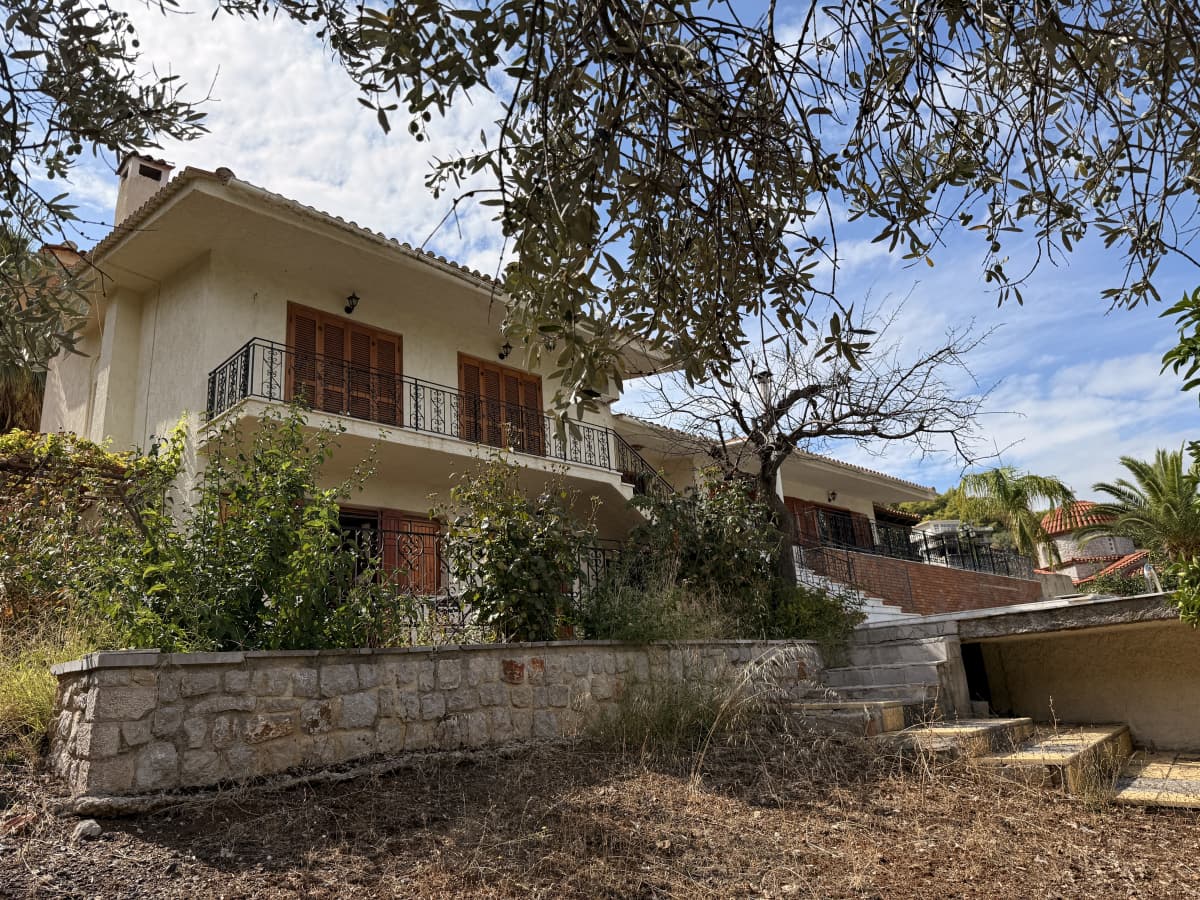 Εξωτερική άποψη διώροφης βίλας με μπαλκόνια, ξύλινα παντζούρια και πέτρινο τοιχίο στο προσκήνιο.