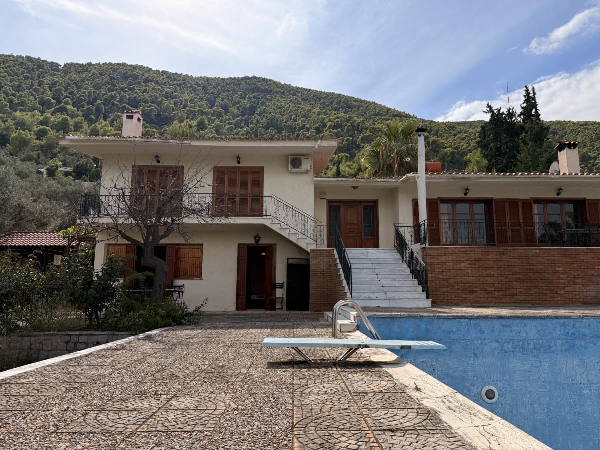 Διώροφο λευκό σπίτι με καφέ πατζούρια και εξωτερική πισίνα μπροστά από καταπράσινο βουνό.