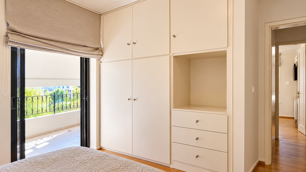 Bedroom featuring a built-in off-white wardrobe with cabinets, drawers, and open shelving next to a sliding glass door with a fabric roman blind opening to a balcony.
