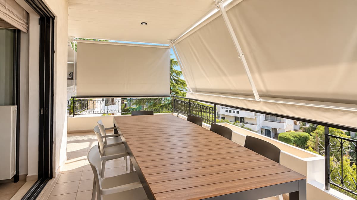 A balcony featuring a long wooden dining table, black chairs, beige awnings, and a metal railing.