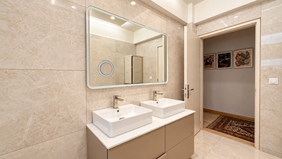 A bathroom features two white vessel sinks on a beige vanity cabinet, a large rectangular mirror with a circular magnifying insert on the tiled wall, and an open door leading to an adjacent room.