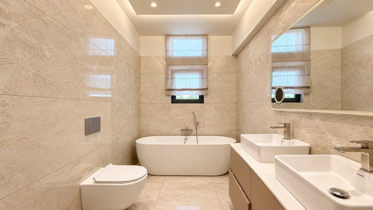 A modern bathroom features beige marble-look tiled walls, a white freestanding bathtub under a window, a wall-mounted toilet, and a double vanity with vessel sinks and a large mirror.