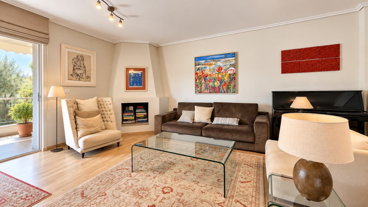 A well-lit living room features a brown sofa, a glass coffee table, a cream armchair, a piano, and various framed artworks on the walls.