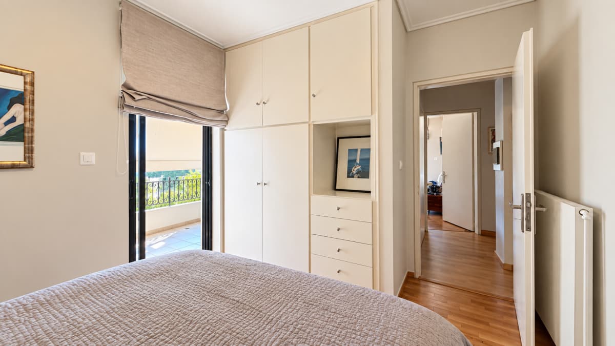 A bedroom features a made bed in the foreground, a built-in off-white wardrobe system with drawers and a small open shelf displaying artwork, and a doorway leading to a hallway.