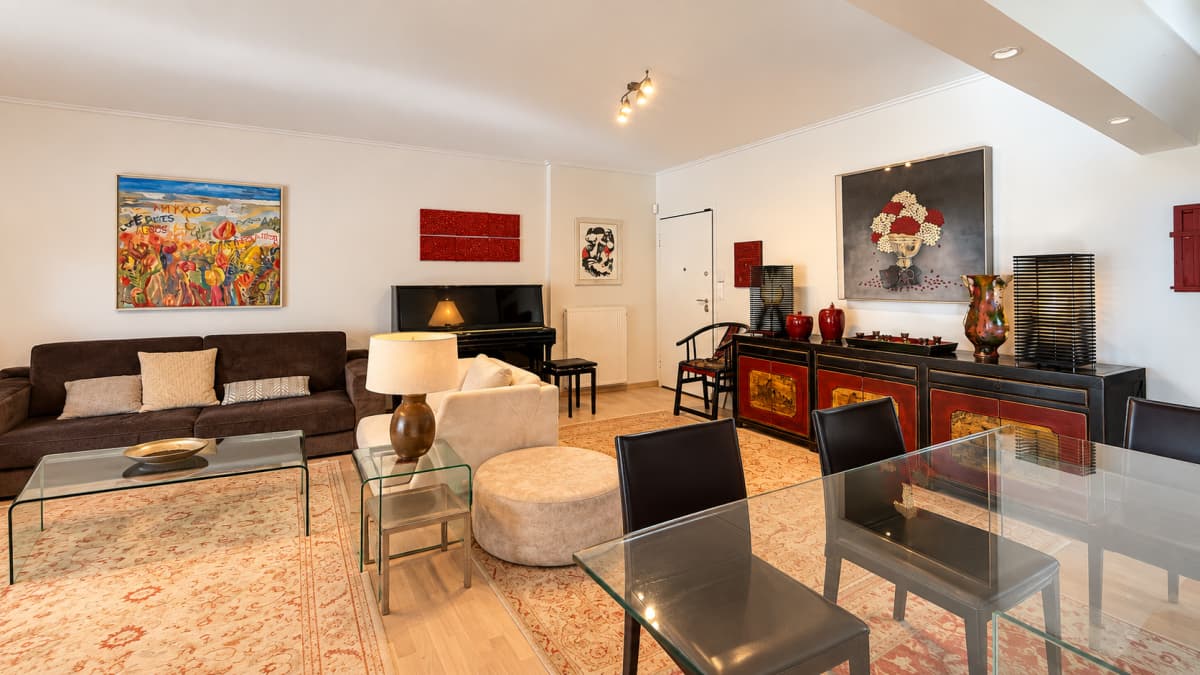 A brightly lit living and dining room features a brown sofa, a piano, a glass-top dining table with black chairs, and colorful artwork on the walls.