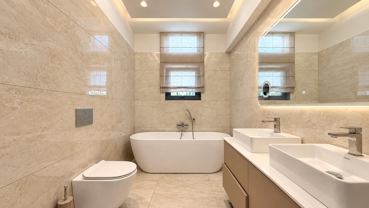 A modern bathroom featuring a white bathtub, dual vessel sinks on a vanity, a wall-mounted toilet, and beige marble-patterned tiles on the walls and floor.