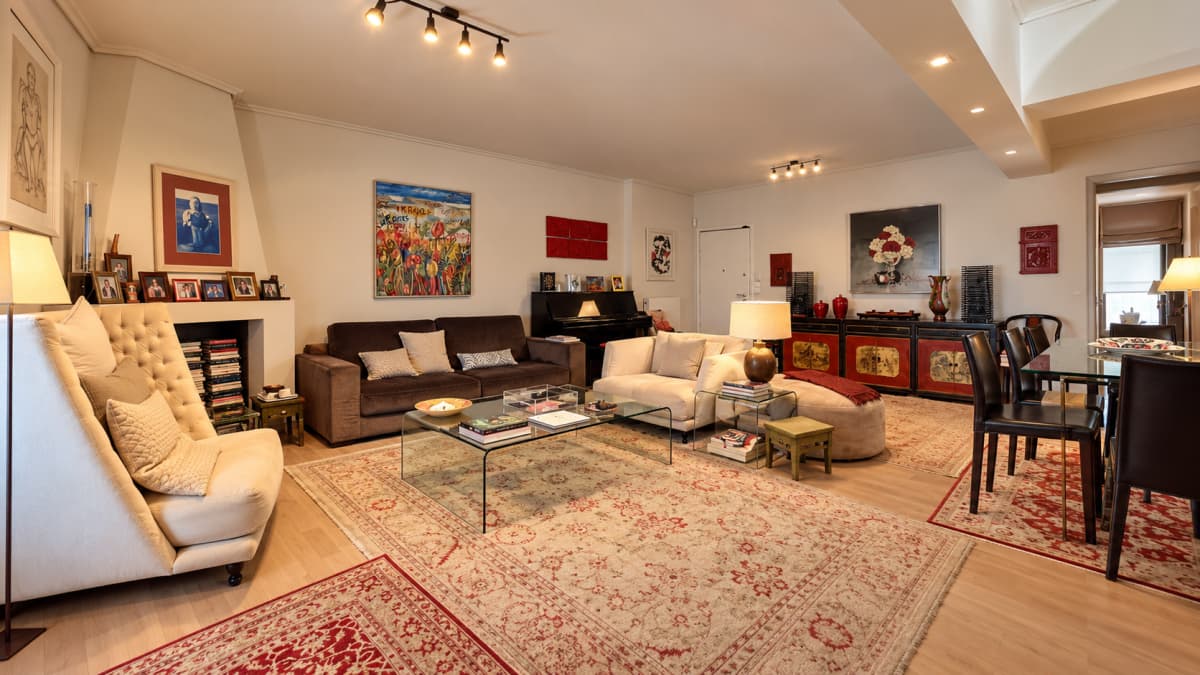 A living room features a cream-colored high-backed armchair, a brown sofa, a white sectional sofa, a glass coffee table, and multiple rugs on a wooden floor.