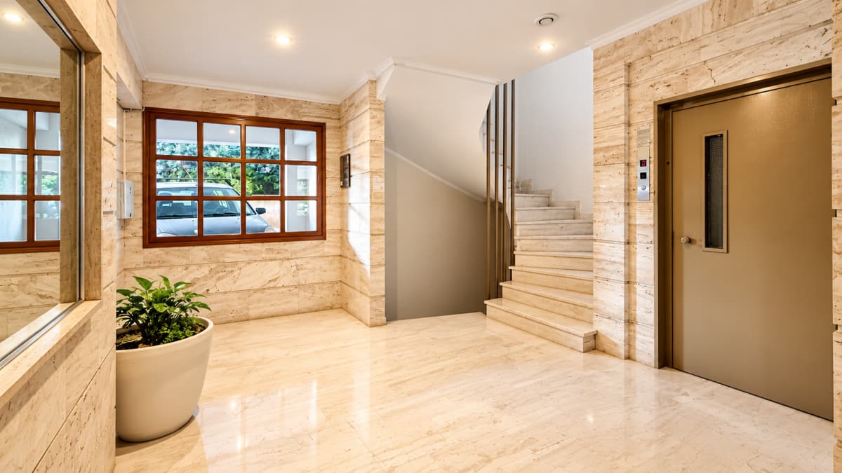 Indoor hallway with travertine walls, polished stone floor, potted plant, window, staircase, and elevator door.