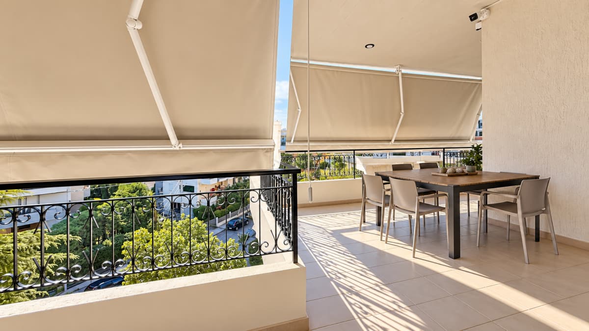 A wide balcony features a dining table with chairs, metal railings, tiled flooring, and sun shades extended overhead.
