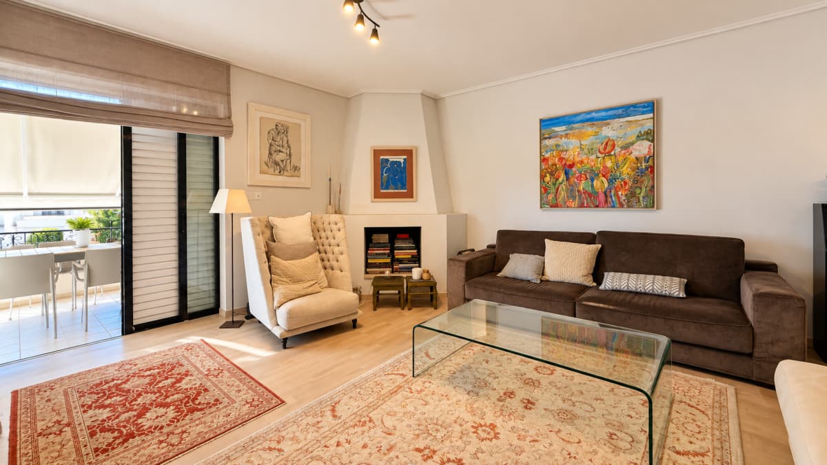 A brightly lit living room features a brown sofa, a glass coffee table, an armchair, a corner fireplace, and two rugs.