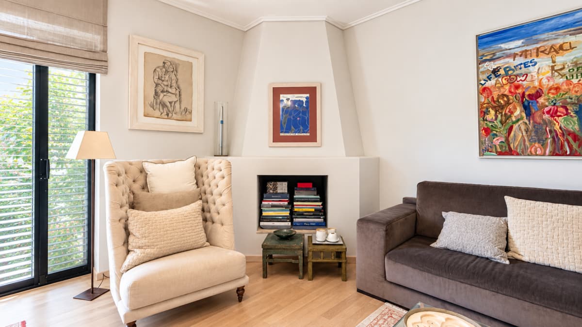 A modern living room features a cream-colored tufted armchair, a dark gray sofa, a corner fireplace filled with books, and art pieces on the white walls.