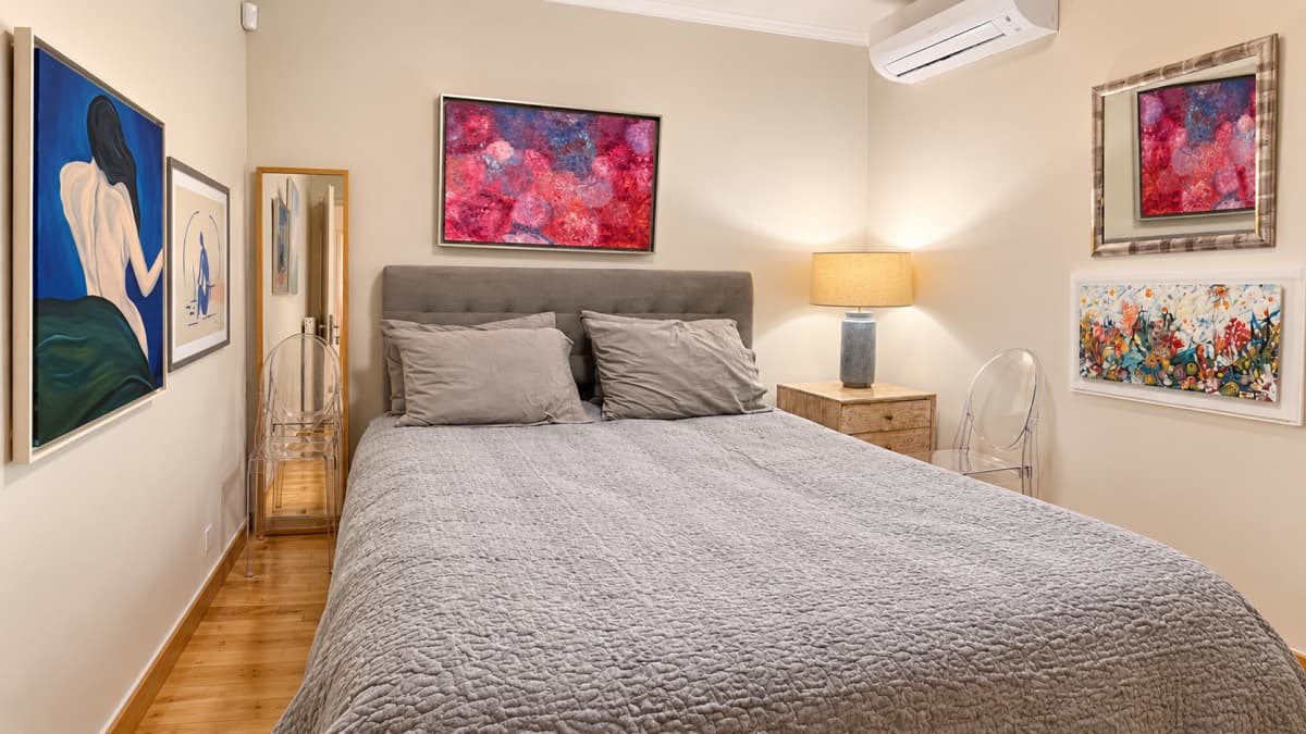 A bedroom features a central grey upholstered bed with two pillows and a grey quilted coverlet, flanked by art pieces, transparent chairs, and a small bedside table with a lamp.