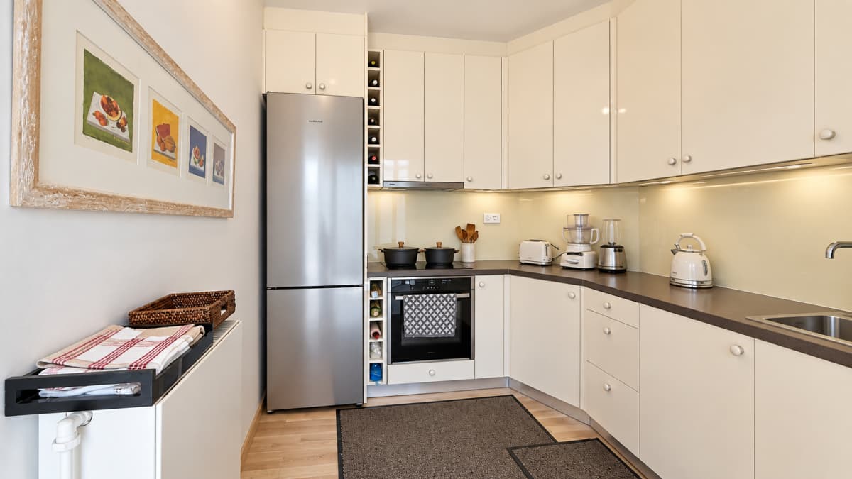 A compact kitchen features cream-colored cabinetry, a stainless steel refrigerator, a built-in oven, and a brown countertop with a sink.