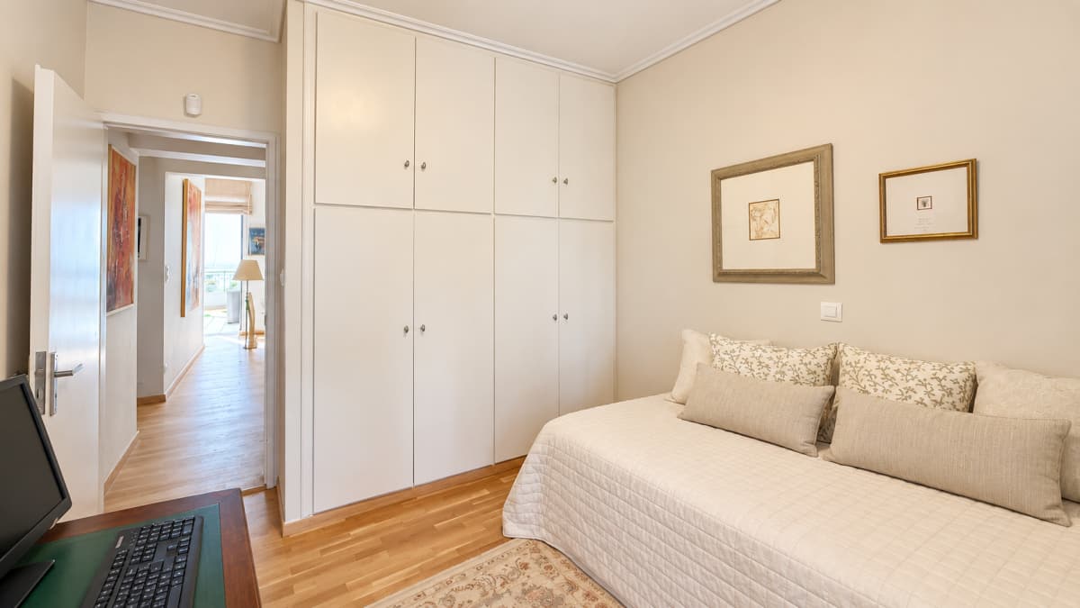 A compact bedroom with a white built-in wardrobe, a single bed with beige pillows, and a wooden desk setup visible in the foreground.