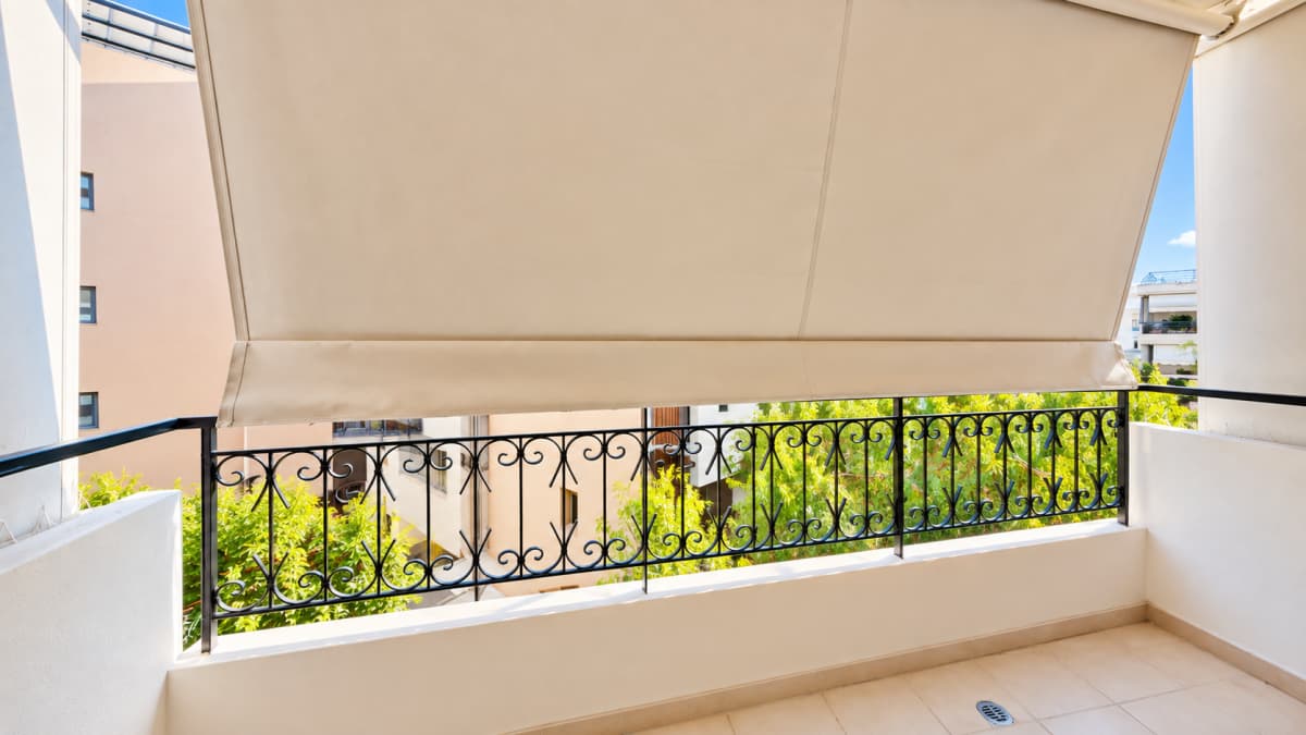 An outdoor balcony with beige tiled flooring, a white low wall, a black decorative metal railing, and a large beige awning extended overhead.