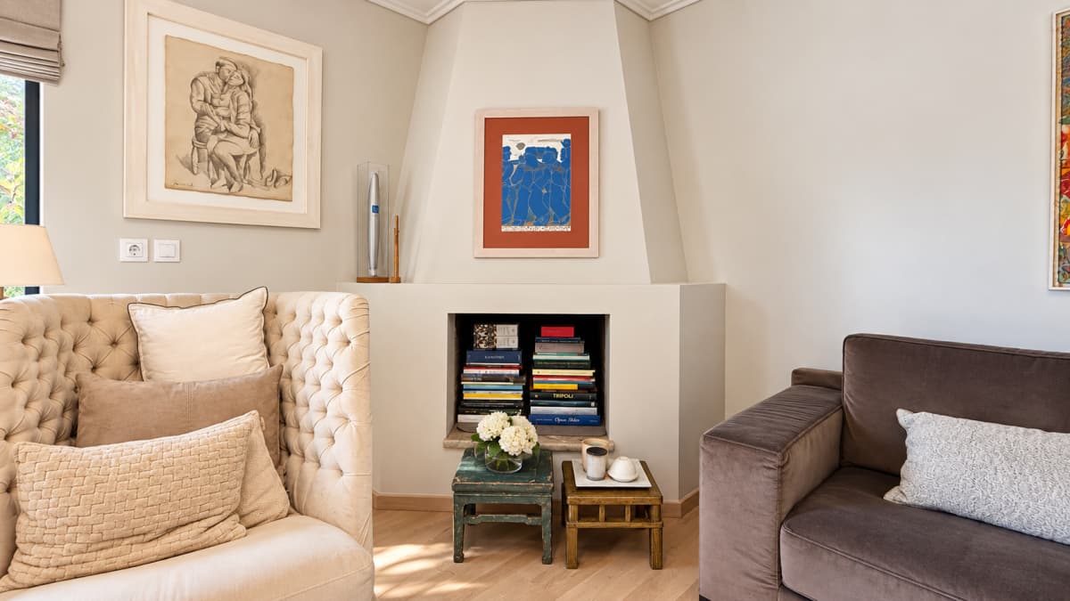 A corner living room featuring a cream tufted armchair, a brown velvet sofa, and a built-in fireplace filled with stacked books.