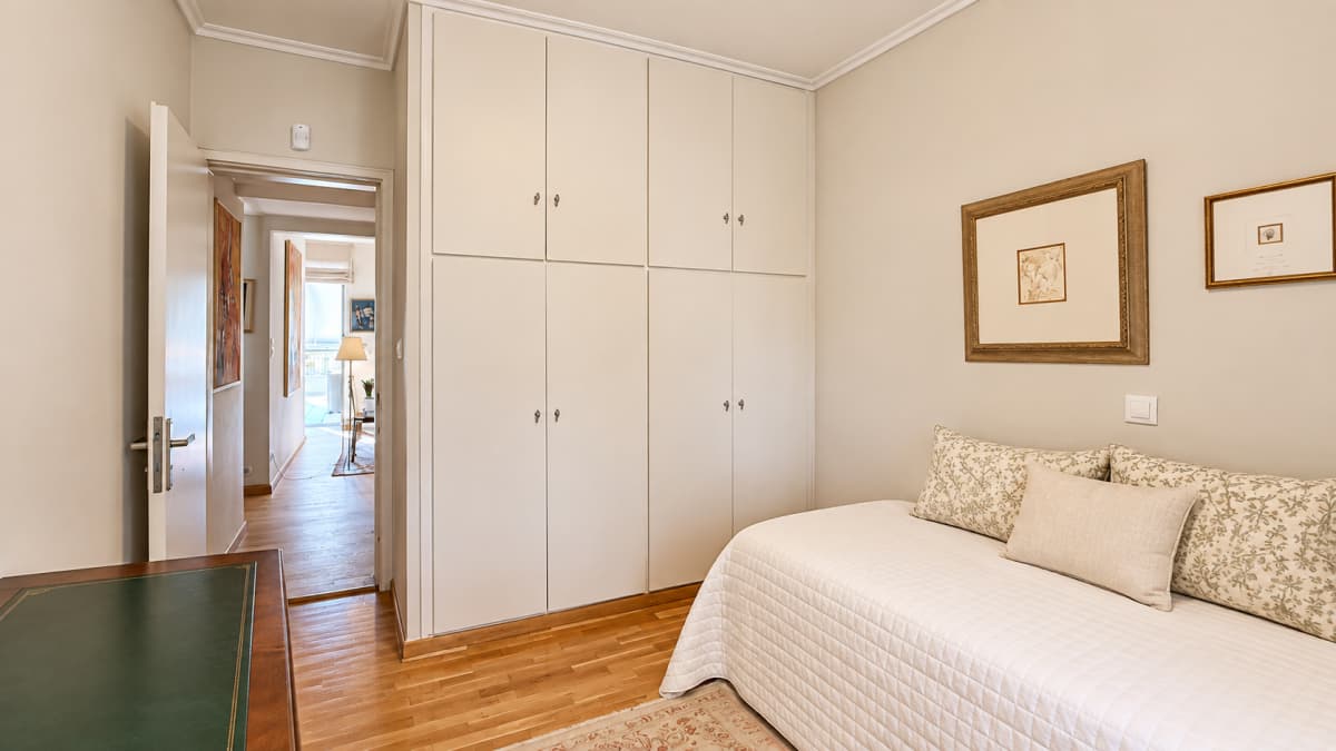 A bedroom features a single bed, built-in wardrobe, framed artwork on the wall, and an open doorway leading to a hallway.