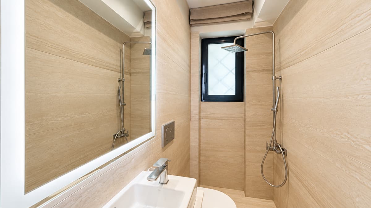 A compact modern bathroom features beige tiled walls, a lit rectangular mirror over a small white sink, and a shower column with an overhead fixture next to a small frosted window.