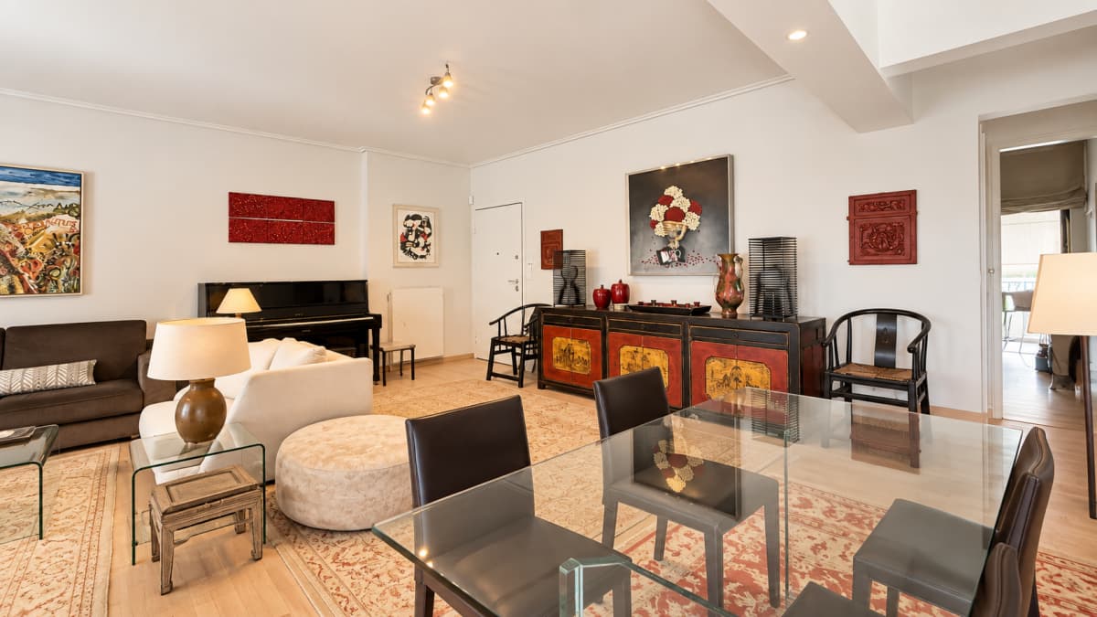 A brightly lit living and dining room features a white sofa, a black upright piano, a glass-topped dining table with black chairs, and Asian-style red and black cabinets decorated with artwork.