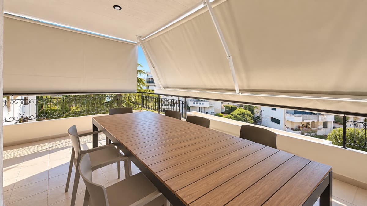 An open balcony features a long wooden outdoor dining table with grey chairs, shaded by beige retractable awnings and surrounded by a black metal railing overlooking buildings.
