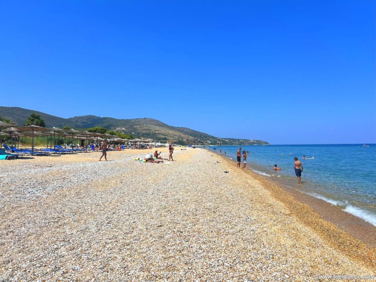 Ευρεία παραλία με βότσαλα και θάλασσα κάτω από καθαρό μπλε ουρανό με βουνά στο βάθος.