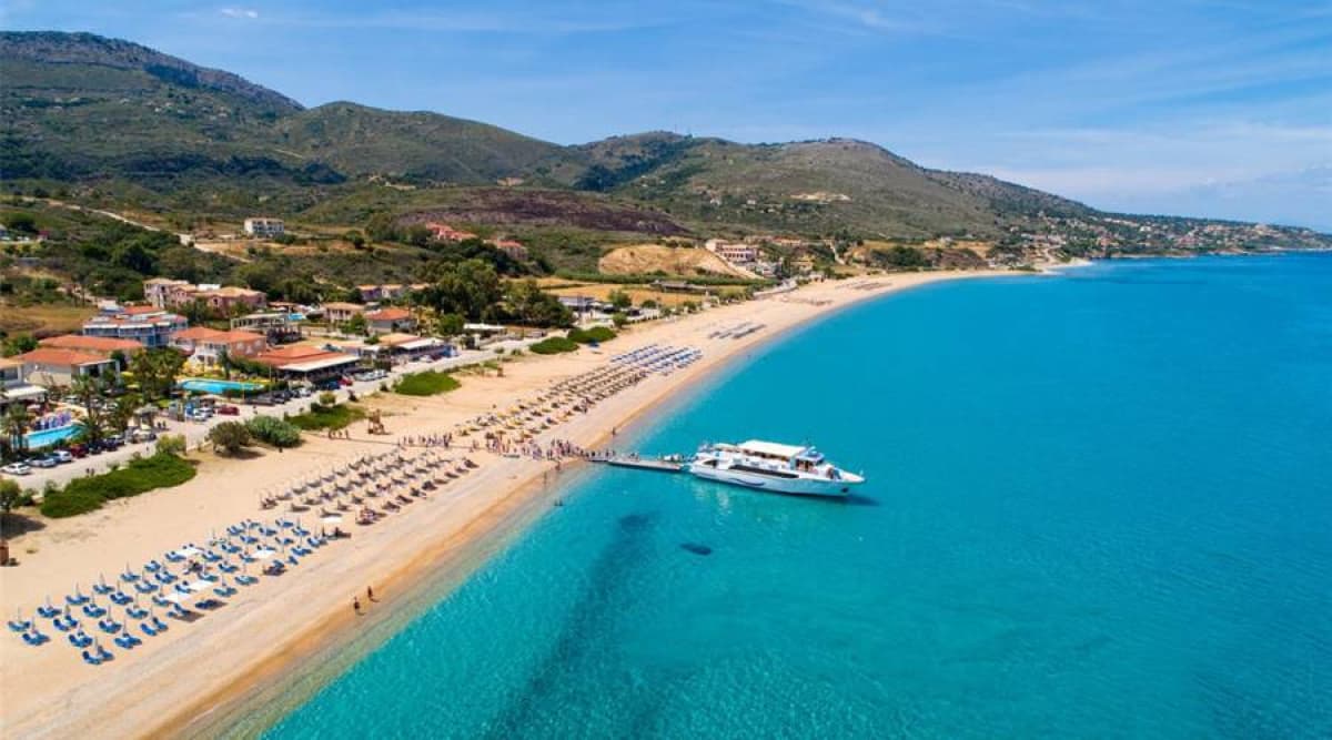 Εναέρια άποψη αμμώδους παραλίας με ξαπλώστρες και ομπρέλες δίπλα σε γαalάζια θάλασσα, με πλοίο δεμένο σε ξύλινη προβλήτα.