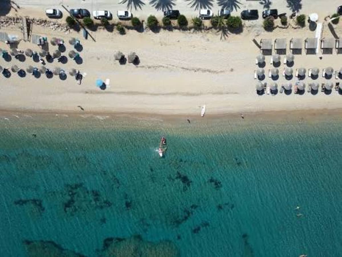 Εναέρια άποψη αμμώδους παραλίας με αυτοκίνητα στον δρόμο, ξαπλώστρες και γαλαζοπράσινα νερά.