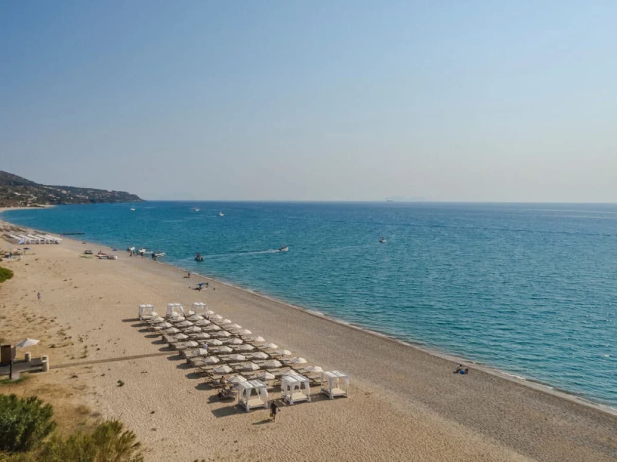 Εναerial άποψη μιας φαρδιάς αμμουδιάς με οργανωμένες ξαπλώστρες και λευκά κρεβάτια με ουρανό κατά μήκος της γαλάζιας θάλασσας.