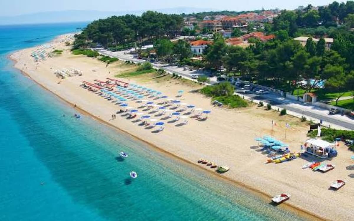 Εναëιά φωτογραφία αμμώδους παραλίας με ομπρέλες και ξαπλώστρες δίπλα σε γαλαζοπράσινα νερά και κτίρια στο βάθος.