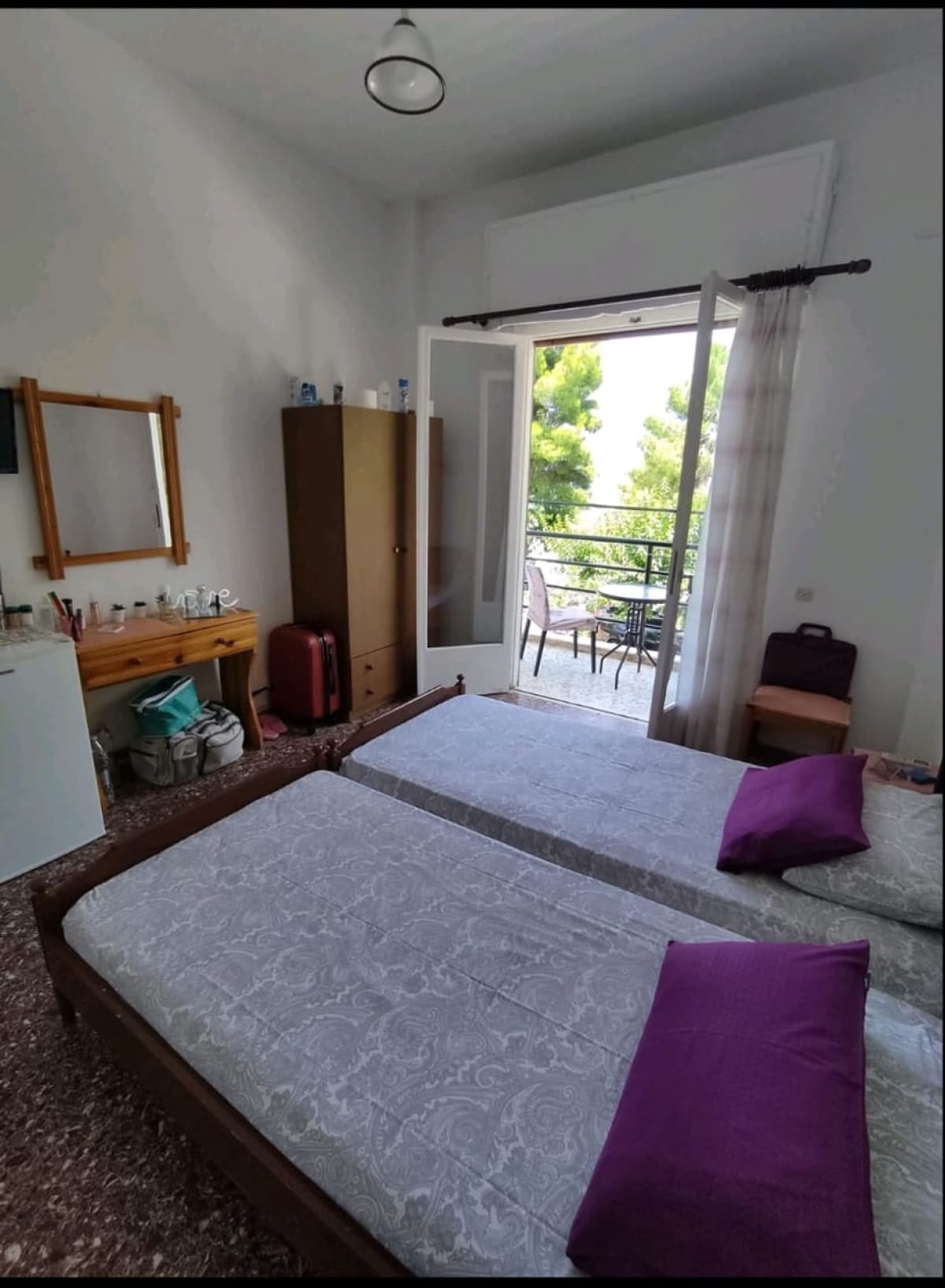 Υπνοδωμάτιο με δύο μονόκλινα κρεβάτια, ξύλινη ντουλάπα, τουαλέτα με καθρέφτη και ανοιχτή μπαλκονόπορτα που βλέπει σε δέντρα.