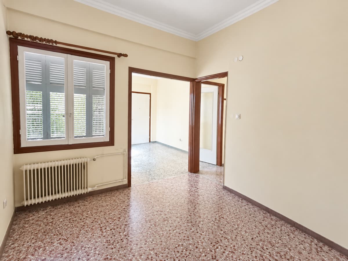 An empty room with beige walls, patterned terrazzo flooring, a window with louvered shutters and a radiator below, and two open doorways leading to another room and hallway.
