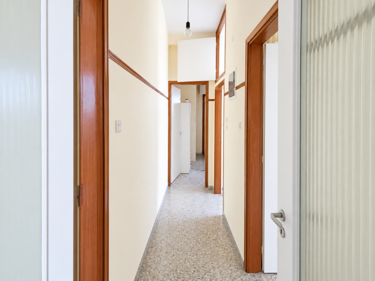 Narrow corridor with cream-colored walls, terrazzo tile flooring, wooden door frames, and multiple doors leading to other rooms.