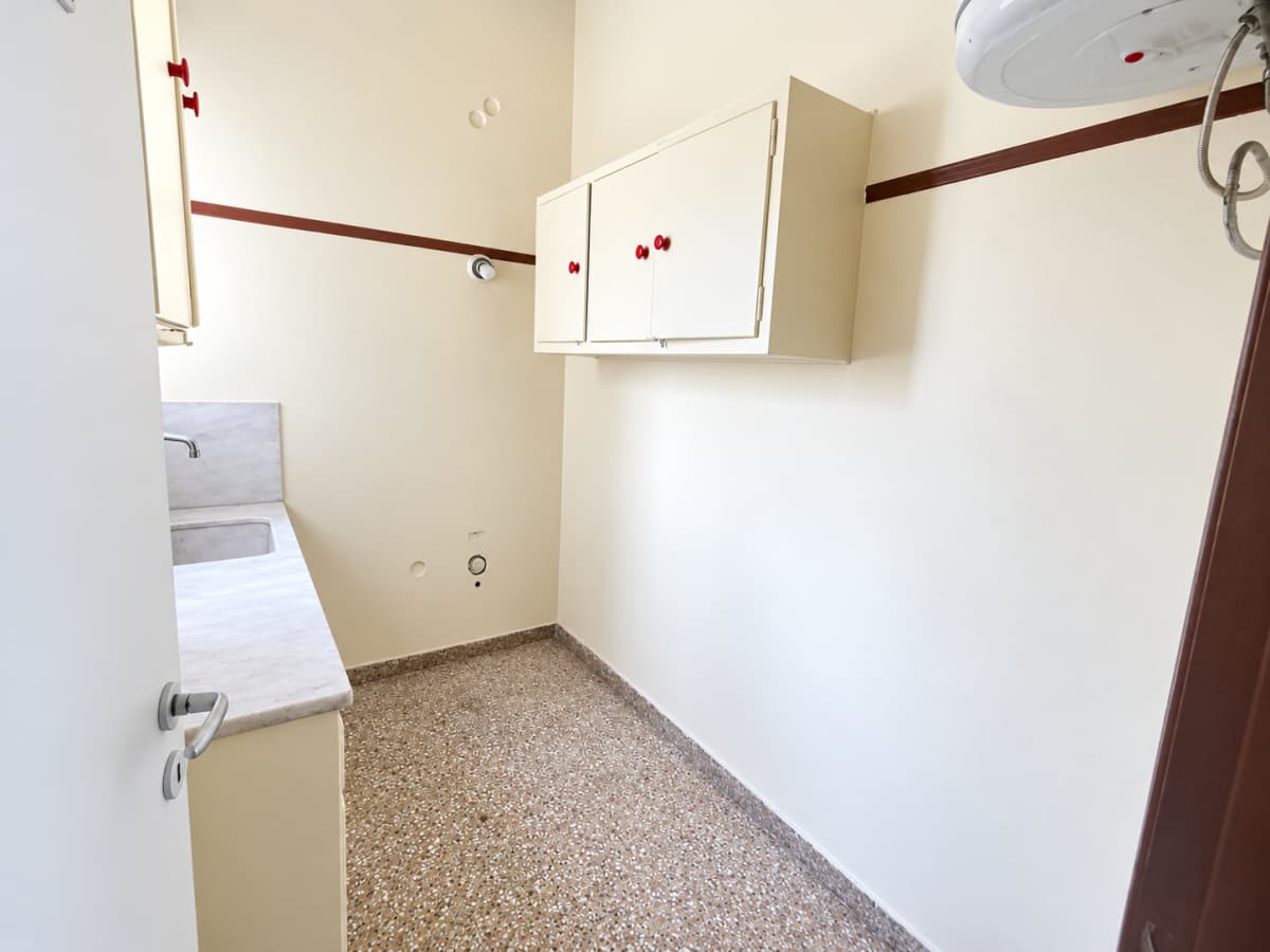 A narrow kitchen or utility room with a counter, sink, wall cabinets, speckled flooring, and a water heater near the ceiling.