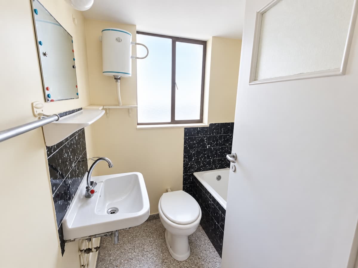 A compact bathroom featuring a white sink, toilet, bathtub, water heater, and a mirror against a pale yellow wall.