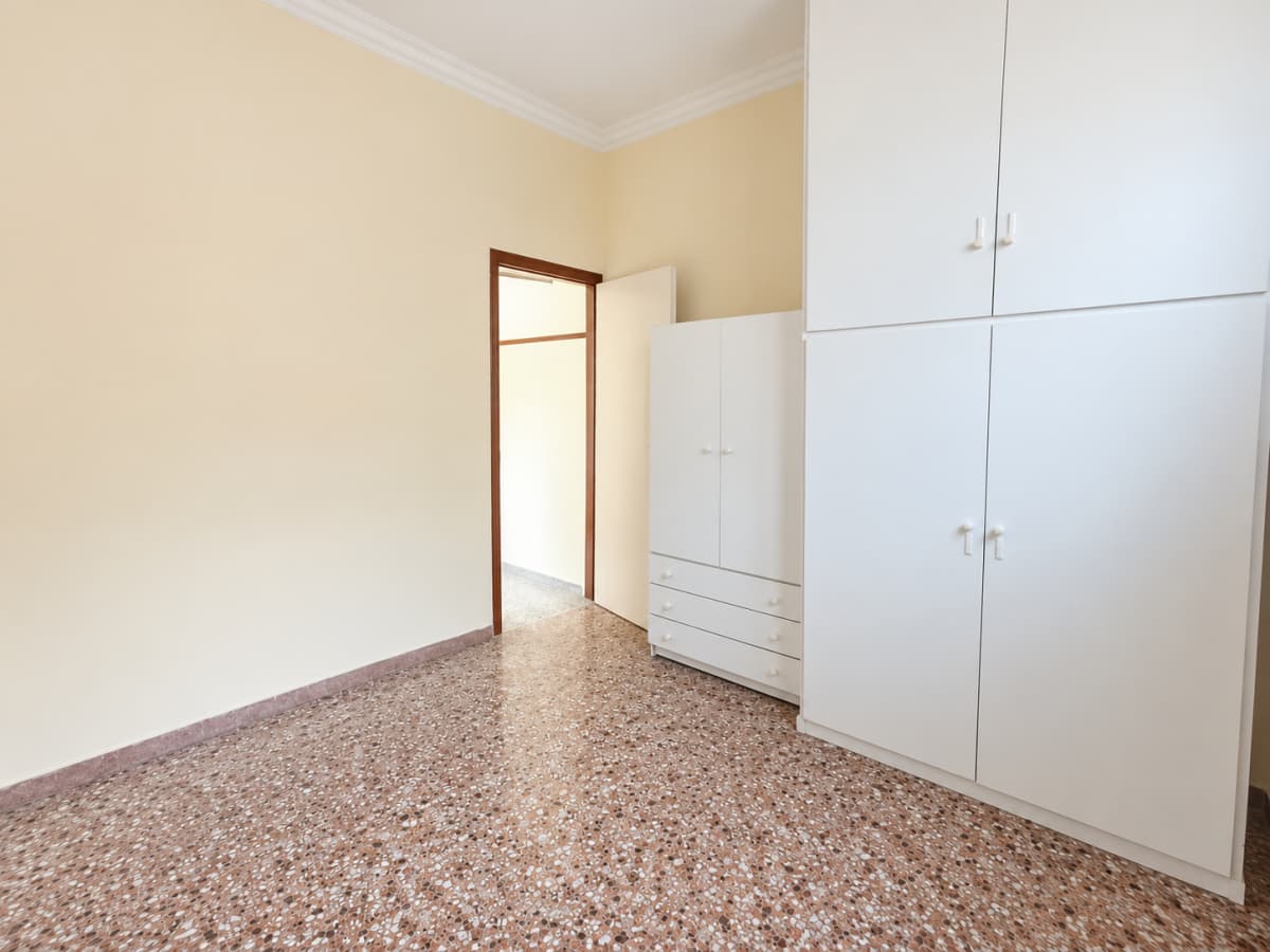 An empty room with beige walls, a speckled terrazzo floor, a built-in white wardrobe on the right, and an open doorway leading to another room.