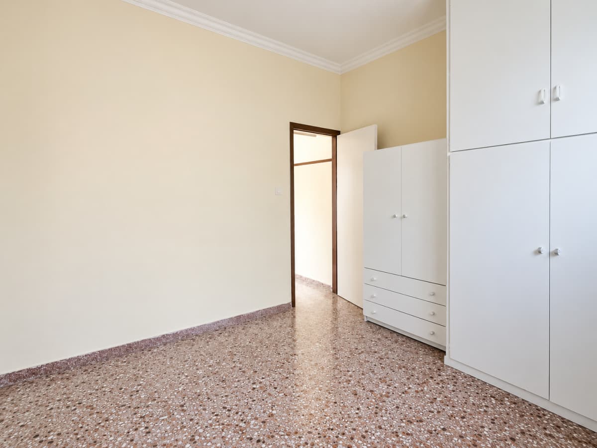 An empty room with beige walls, a speckled tile floor, white built-in wardrobes, and an open doorway leading to another hallway.