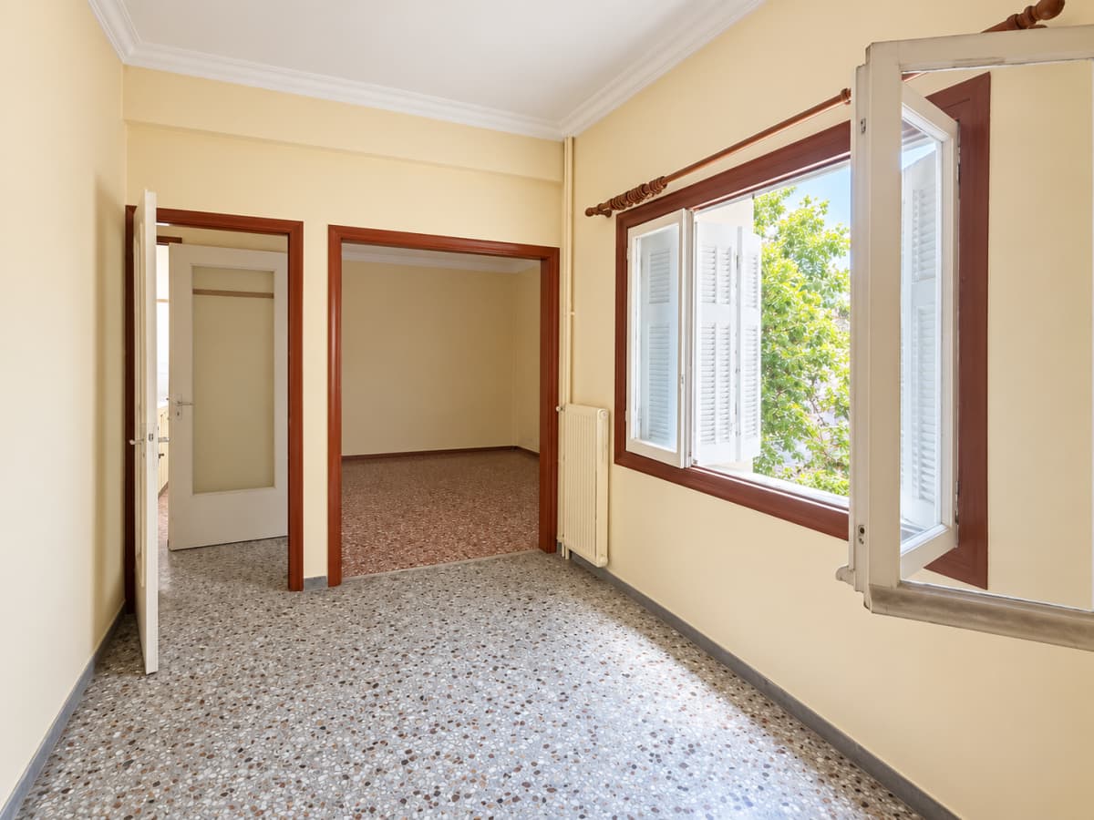 An empty room with terrazzo flooring, pale yellow walls, two doorways, and an open window with a view of greenery.