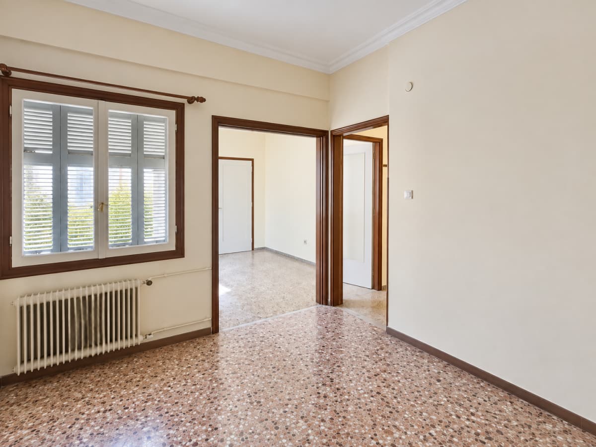 An empty room with terrazzo flooring, off-white walls, a window with louvers and a radiator below, and two doorways leading to other rooms.