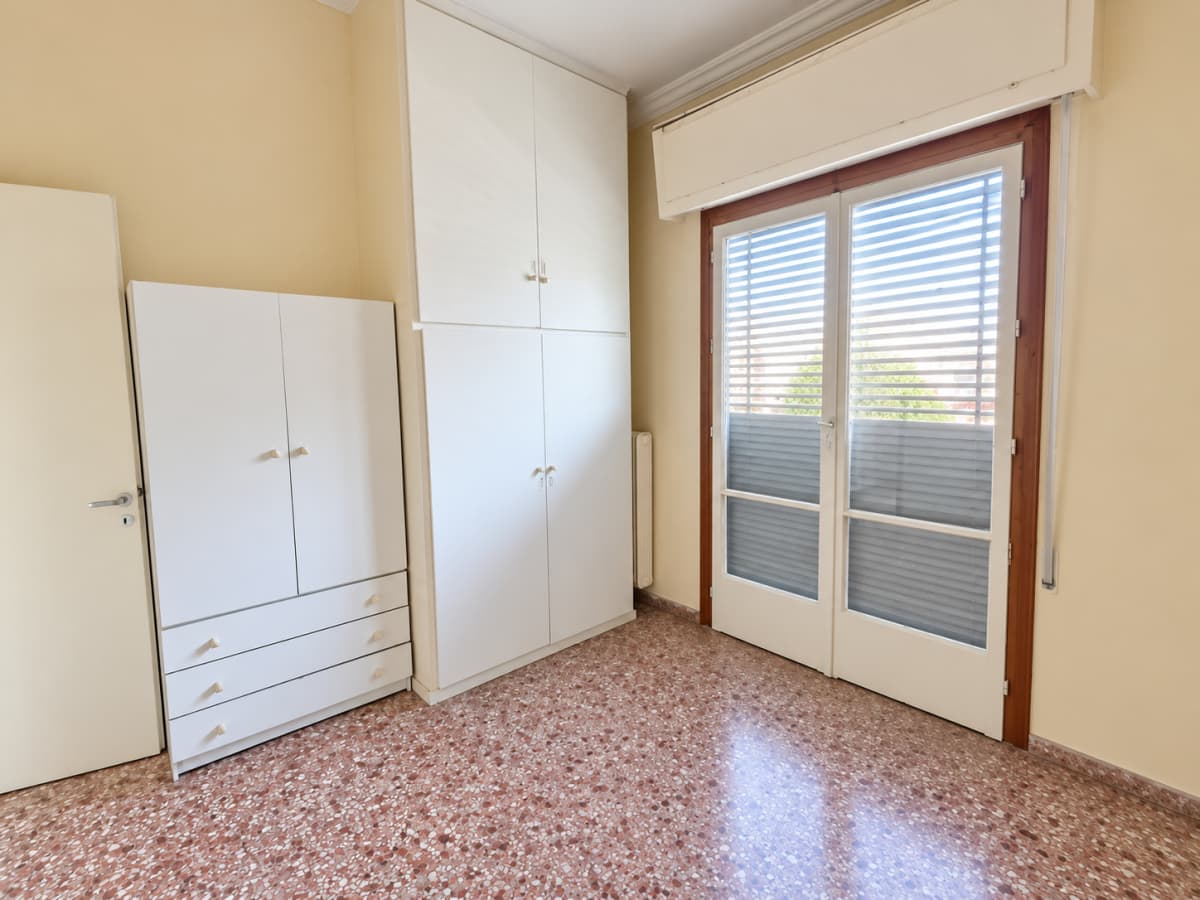 Interior view of a bedroom featuring two white wardrobes, light yellow walls, patterned tile flooring, and a large window with blinds.