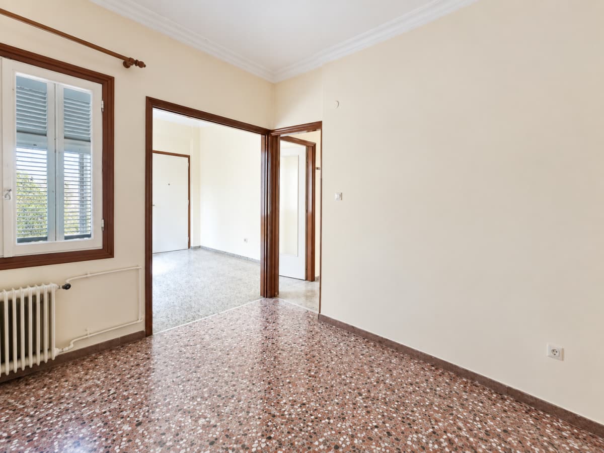 Interior room with reddish terrazzo flooring, cream-colored walls, a window with brown wooden shutters on the left, a radiator below the window, and two open doorways leading to another room and hallway.