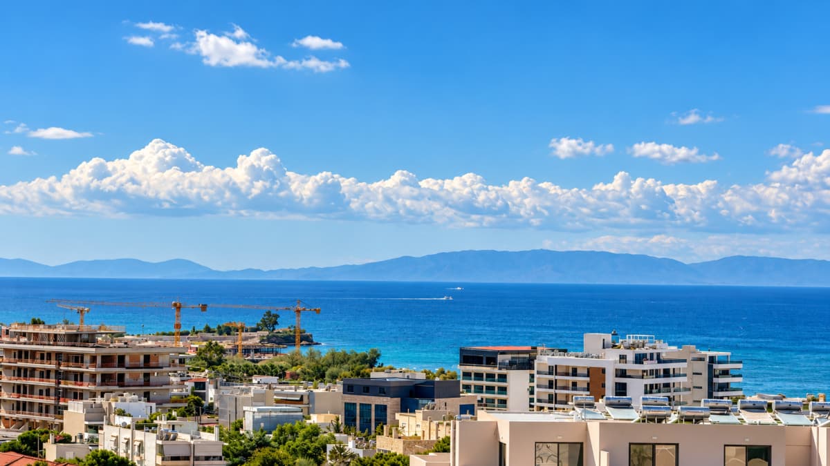 Εξωτερική λήψη από ψηλά δείχνει παράκτια κτίρια και γερανούς εργοταξίου με φόντο τη θάλασσα και βουνά κάτω από συννεφιασμένο ουρανό.