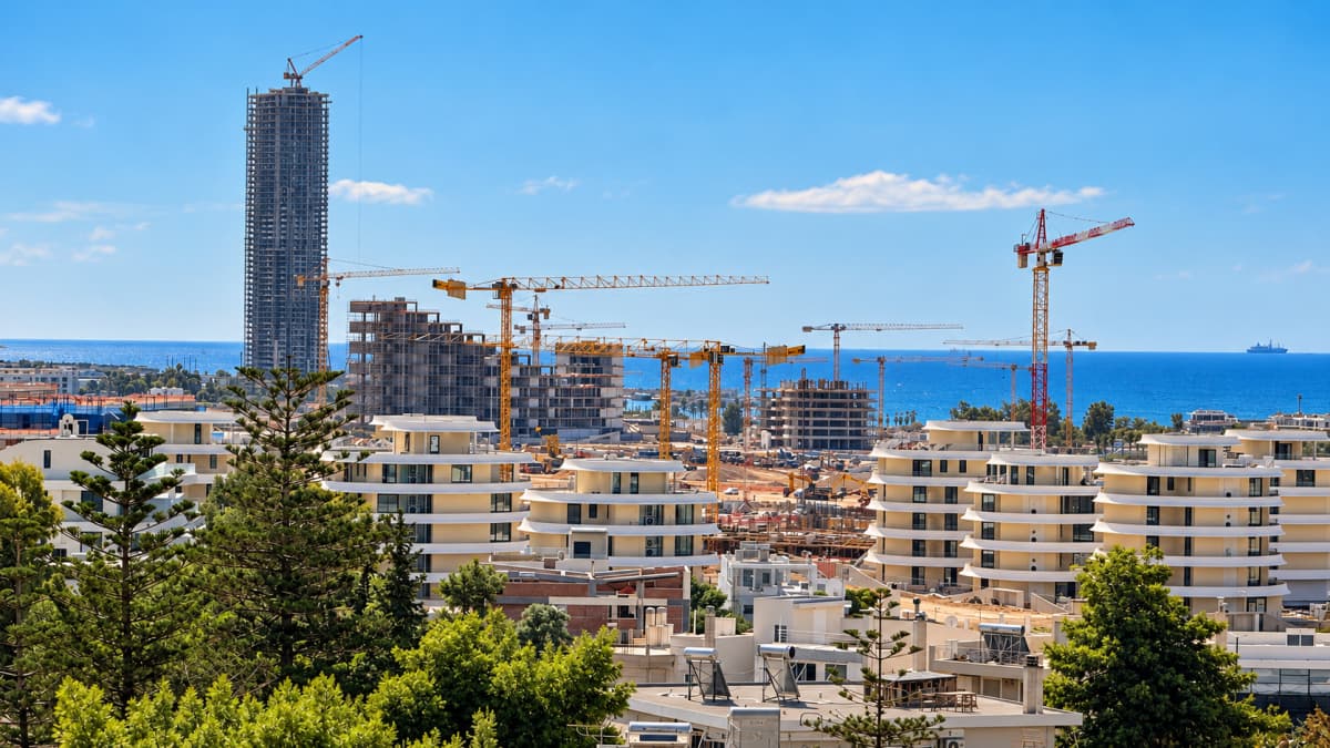 Εξωτερική άποψη εργοταξίου με γερανούς και πολυκατοικίες μπροστά από τη θάλασσα.